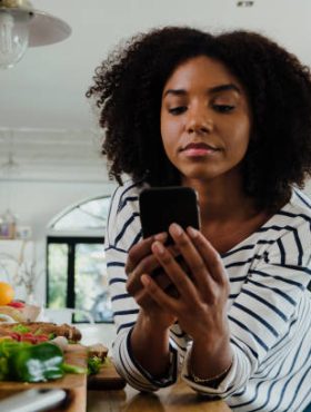 Beautiful focussed African American female researching lunch recipe on smartphone standing next to chopped vegetables in organised kitchen. High quality photo